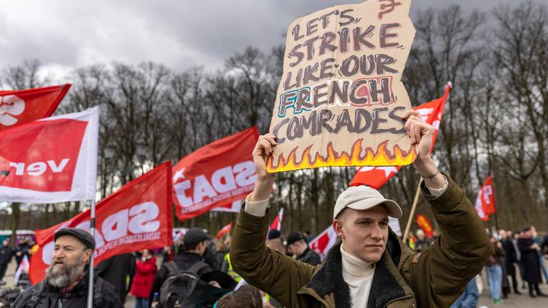 Mehrere Menschen nehmen am 25. März 2023 an einem Gewerkschaftsprotest teil. Ein Mann hält ein Schild mit der Aufschrift „ "Lasst uns wie unsere französischen Genossen zuschlagen". Mehrere Menschen nehmen am 25. März 2023 an einem Gewerkschaftsprotest teil. Ein Mann hält ein Schild mit der Aufschrift „ "Lasst uns wie unsere französischen Genossen zuschlagen".